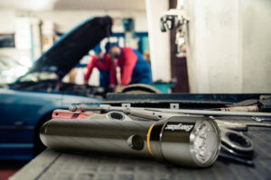 ENR_Vision_HD_6AA_Lifestyle Image_PMZH61_Garage an Energizer Vision HD 1AA Metal Flashlight on the table and 2 mechanic working on a car in background